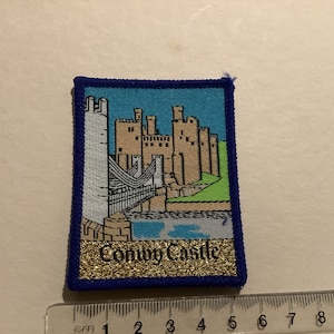Puede incluir: Un parche bordado azul y dorado con una representación del Castillo de Conwy en Gales. El parche tiene un borde azul y el texto "Conwy Castle" en purpurina dorada.