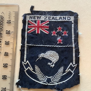 May include: A rectangular patch with the words "NEW ZEALAND" at the top. The patch features the New Zealand flag, four red stars, a kiwi bird, and fern leaves, all in white and red on a navy blue background.