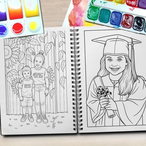 May include: Spiral-bound coloring book with two detailed black and white illustrations. One shows two children in front of sunflowers, the other a girl in a graduation cap and gown. Paintbrushes and watercolor paints are arranged around the book.