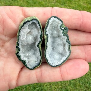 May include: Two small, matching halves of a geode stone featuring a rough, moss-green outer crust and a hollow center filled with sparkling, translucent white quartz crystals. The stones have an irregular, oval shape with dark green edges.