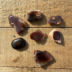 May include: A collection of polished agate stones in shades of brown, red, and cream. The stones vary in shape and size, with some displaying banded patterns and others featuring unique inclusions. The stones are arranged on a weathered wooden surface.