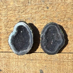 May include: Two halves of a geode, each with a dark, sparkling interior and a contrasting light-colored border. The geodes are displayed on a wooden surface, showcasing their natural beauty and unique crystalline structures.
