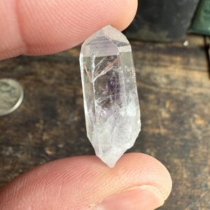 May include: A clear quartz crystal point, held between fingers. The crystal is transparent with internal fractures and a faceted top. The crystal is approximately 1.5 inches long, with a rough, natural base.