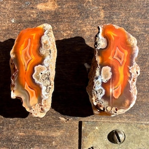 May include: Two polished agate slices with vibrant orange, yellow, and white banding. The natural stone pieces have a rough, textured exterior and are displayed on a wooden surface. The agate's patterns create a striking visual contrast.