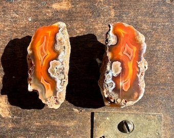 Puede incluir: Dos rodajas de ágata pulidas con bandas vibrantes de color naranja, amarillo y blanco. Las piezas de piedra natural tienen un exterior rugoso y texturizado y se exhiben sobre una superficie de madera. Los patrones de la ágata crean un contraste visual llamativo.
