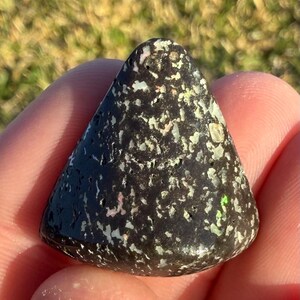 May include: A dark, triangular-shaped stone with a speckled pattern of white and iridescent flecks. The stone has a smooth, polished surface and is held in a person's hand. The stone's color is primarily black with hints of green and pink.