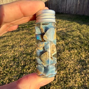 May include: A small glass vial filled with blue and tan gemstones. The vial has a silver metal screw-on cap. The gemstones are a mix of light blue and tan colors. The vial is held in a person's hand.