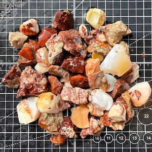 May include: A collection of raw, unpolished stones in shades of red, orange, cream, and white, scattered across a black cutting mat with white grid lines and numerical markings, showcasing varied textures, jagged edges, and smooth, translucent surfaces.