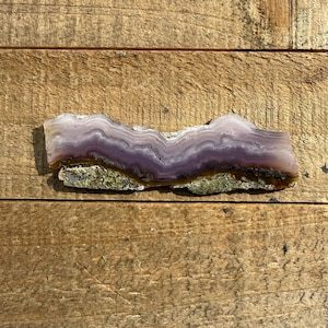 May include: A polished, natural stone slab with a layered, wavy pattern. The stone features bands of lavender and white, with a rough, mossy base. The stone is displayed on a rustic, wooden surface.