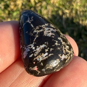 May include: A polished, black gemstone with iridescent white and pink inclusions. The stone has a smooth, rounded shape and is held in a hand against a blurred green background. The surface of the stone reflects light.