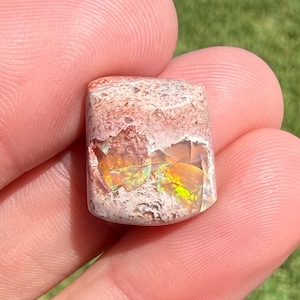 May include: A rectangular gemstone with a mix of colors, including white, brown, and iridescent flashes of yellow and green. The stone is held between fingers, showcasing its natural beauty and unique patterns. The gemstone is a unique piece of jewelry.