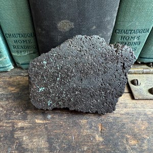 May include: A dark, textured, rectangular object with turquoise accents rests on a wooden surface. Behind it are several vintage green books with the text "Chautauqua Home Reading Series." A metal hinge is visible.