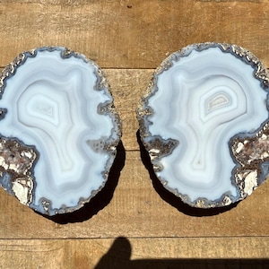 May include: Two agate slices with a white and blue banded pattern. The agate slices have a natural, rough edge and are set against a wooden background.