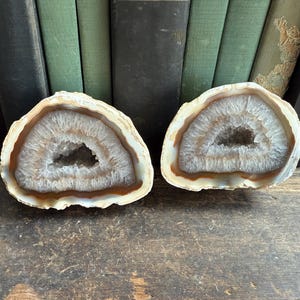 May include: Two polished agate geodes, each cut in half to reveal a crystalline interior. The outer edges are a creamy white, transitioning to a band of brown and a central cavity filled with sparkling crystals. The background includes vintage books.
