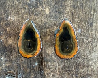 May include: Two matching agate stone pendants with a teardrop shape. The stones feature a dark center, surrounded by bands of orange, yellow, and green, with a silver-colored edge. The pendants are displayed on a wooden surface.