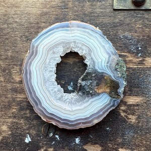 May include: A white and brown agate slice with a hole in the center. The agate has a natural, organic shape and is polished to a smooth finish.
