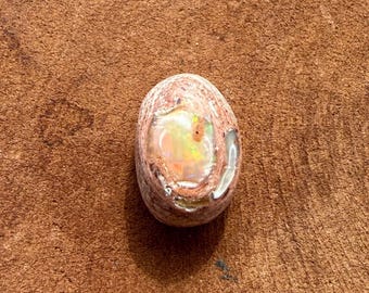 May include: An oval-shaped opal gemstone, polished to a smooth finish. The opal displays a play of colour, with flashes of yellow, orange, and green in its translucent core. The outer layer is a mix of tan and brown.