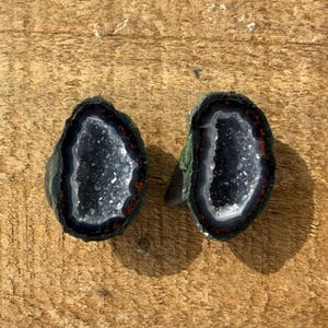 May include: Two polished agate geodes with dark gray and white crystal interiors, framed by a black, white, and red border. The geodes are displayed on a textured, light brown wooden surface.