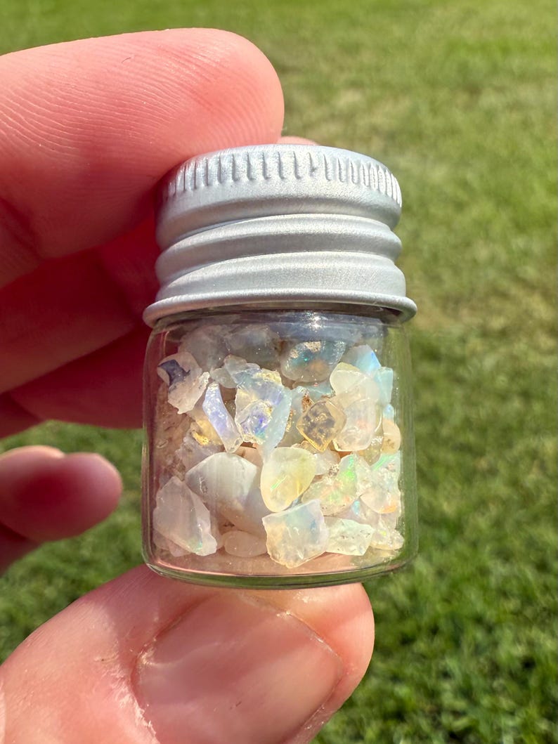 Pu&ograve; includere: Un piccolo barattolo di vetro pieno di frammenti di opale iridescenti. Il barattolo ha un coperchio a vite in metallo argentato. Gli opali presentano sfumature bianche, gialle e blu. Il barattolo &egrave; tenuto in mano, su uno sfondo di erba verde.