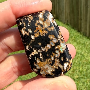 May include: A polished, rectangular stone with a black base and intricate patterns of white, gold, and iridescent colors. The stone is held in a hand, showcasing its smooth, reflective surface and natural beauty.