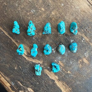 May include: A collection of turquoise gemstones with a vibrant blue-green hue and dark matrix patterns. The stones vary in size and shape, arranged on a rustic, weathered wooden surface. The natural gemstones are unpolished and have a raw, organic appearance.