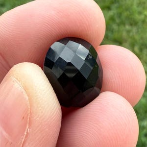 May include: A faceted, oval-shaped black gemstone is held between fingers. The gem has a glossy, reflective surface with multiple geometric facets. The background is a blurred view of green grass.