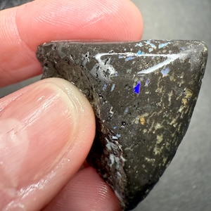 May include: A triangular-shaped, rough-textured stone with a dark gray base color. The stone has iridescent blue and white flecks throughout, with a prominent blue flash near the top. The stone is held between two fingers.
