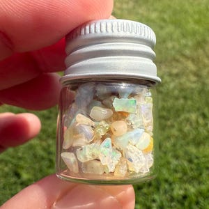 Pu&ograve; includere: Un piccolo flacone di vetro pieno di schegge di opale iridescenti. Il flacone ha un tappo in metallo argentato ed &egrave; tenuto dalle dita di una persona. Gli opali mostrano una gamma di colori, tra cui bianco, giallo e verde, su uno sfondo verde sfocato.