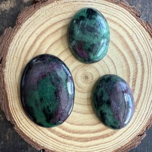 May include: Three polished oval gemstones with a mix of green, dark purple, and red hues. The stones are displayed on a round, light brown wooden slice with visible grain patterns. The gemstones have a glossy finish.
