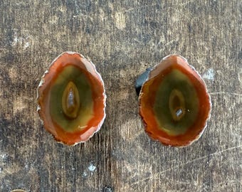 May include: Two oval-shaped agate stones with a polished surface. The stones feature a central olive green band, surrounded by a band of orange-red, and a silver-colored edge. The stones are displayed on a wooden surface.