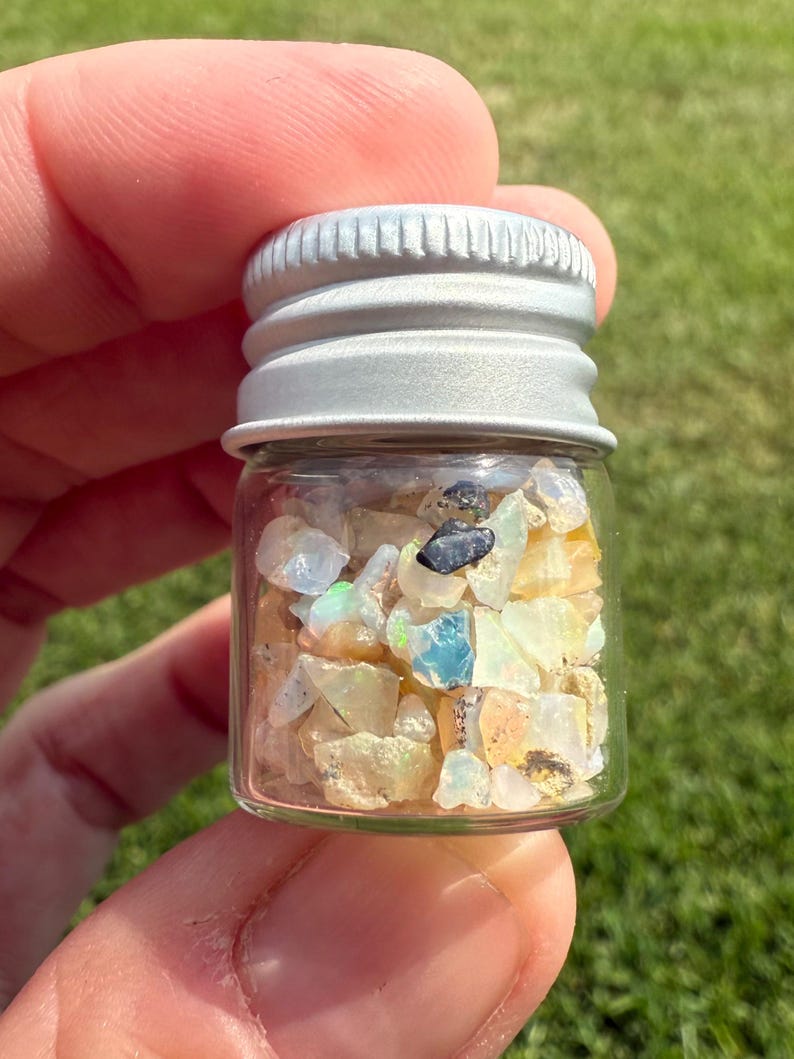 Pu&ograve; includere: Un piccolo flacone di vetro pieno di varie pietre preziose di opale grezze e colorate. Gli opali mostrano una gamma di colori, tra cui bianco, giallo, blu e nero. Il flacone ha un coperchio a vite in metallo argentato. Lo sfondo &egrave; erba verde sfocata.