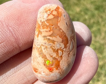 May include: A polished, oval-shaped gemstone featuring a mottled surface with shades of peach, tan, and cream. The stone displays small, vibrant flashes of neon green and bright orange fire within its textured, earthy-toned matrix.
