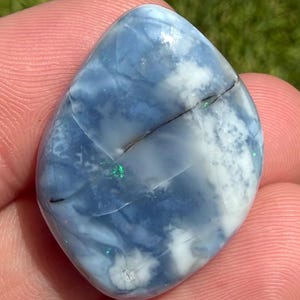 May include: A polished, teardrop-shaped blue opal gemstone featuring swirling patterns of milky white and translucent light blue. The surface displays subtle, dark linear inclusions and small, vibrant flashes of iridescent green fire.