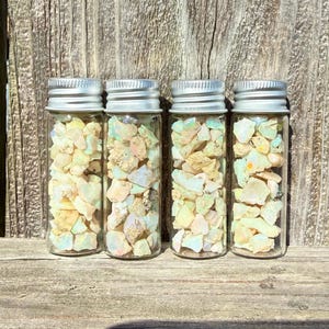 May include: Four clear glass vials with silver-colored screw-top lids, each filled with small, irregular, translucent opal stones displaying iridescent flashes of white, pale yellow, and light green colors throughout the collection.