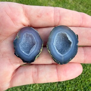May include: Two small, polished agate slices featuring deep blue-grey outer bands and sparkling, crystalline druzy centers. The stones have irregular, rounded shapes with dark, textured edges, showcasing a natural layered mineral formation.