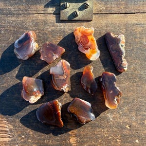 May include: A collection of polished agate stones in shades of brown, orange, and white, arranged on a weathered wooden surface. The stones vary in size and shape, showcasing natural patterns and textures. The background includes a metal plate with screws.