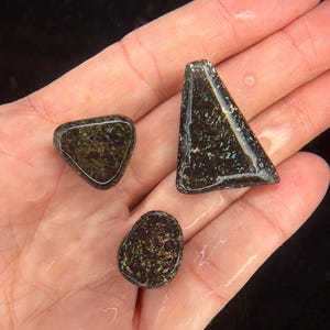 May include: Three polished black opal gemstones in various shapes: a triangle, a rounded triangle, and an oval. The opals display a dark base color with iridescent flecks of green, blue, and gold. The gemstones are wet, reflecting light.