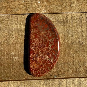 May include: A polished, semi-circular stone with a mottled pattern of deep red, brown, and tan hues. The stone's surface is smooth and glossy, showcasing a natural, organic design. The stone is set against a wooden background.