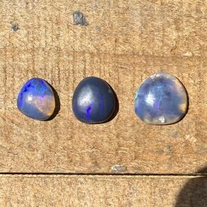 May include: Three smooth, polished, rounded gemstones arranged in a row. The left stone is translucent with blue and orange flashes. The center stone is dark, opaque, and deep blue. The right stone is larger, translucent, and pale blue.