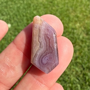 May include: A polished, irregularly shaped stone with a swirling pattern of lavender, tan, and white. The stone has a smooth, glossy surface and is held against a backdrop of green grass. The stone's colors and patterns create a unique, natural aesthetic.