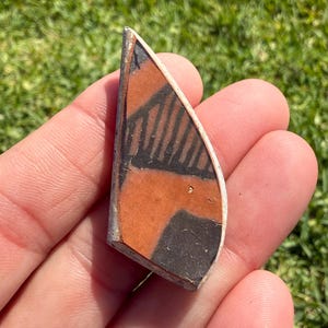 May include: A fragment of pottery with a geometric design. The shard features a combination of black, orange, and white colors. The design includes black lines and shapes on an orange background, with a white border.
