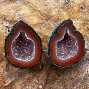 May include: Two matching halves of a geode featuring a dark green outer rind, a thick band of deep reddish-brown agate, and a central cavity filled with sparkling white crystalline structures. The stones are teardrop-shaped with smooth edges.