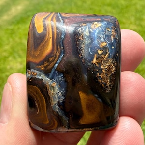May include: A polished rectangular gemstone featuring swirling patterns of deep brown, golden amber, and black, accented by a translucent blue vein and embedded metallic gold-colored flecks throughout the smooth, reflective surface of the stone.