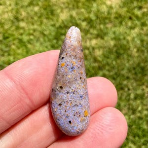 May include: A teardrop-shaped polished stone featuring a light lavender base color with a speckled texture. The surface is covered in small, irregular inclusions of dark brown, black, and bright orange flecks distributed throughout the material.