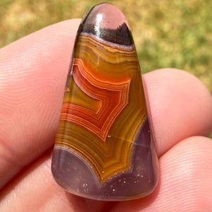 May include: A polished, triangular-shaped agate gemstone featuring distinct, concentric bands of burnt orange, golden yellow, and deep purple. The stone has a smooth, glossy surface with a translucent top section and opaque, layered patterns throughout.