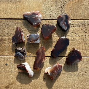 May include: A collection of polished agate stones in shades of brown, red, and white, arranged on a wooden surface. The stones vary in shape and size, showcasing natural patterns and textures. Ideal for collectors or for use in jewelry making.