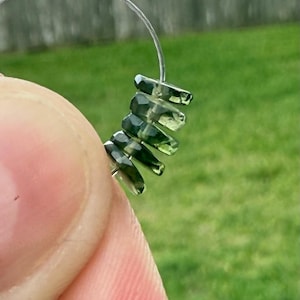 May include: Close-up of a section of a beaded earring. The beads are faceted, dark green, and rectangular. They are strung on a thin, clear wire that forms a loop. The background is blurred green grass and a wooden fence.