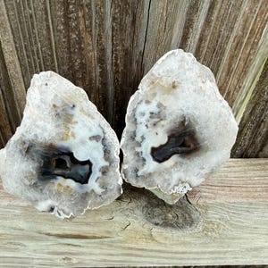 May include: Two halves of a geode stone featuring a crystalline outer edge and a dark, hollow center. The mineral structure displays layers of white, translucent, and grey tones surrounding a deep black cavity within the rock formation.