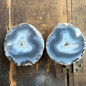 May include: Two circular agate slices featuring concentric bands of light blue, gray, and white. Each stone has a rough, textured outer edge and a small, crystalline hollow center. The stones are positioned side by side on a flat surface.