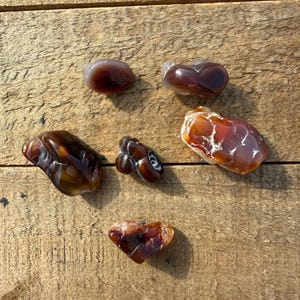May include: A collection of polished agate stones in shades of brown, orange, and white, arranged on a wooden surface. The stones vary in shape and size, with some displaying unique patterns and textures. The natural gemstones are ideal for jewelry making or collecting.